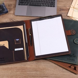 May include: A dark brown leather portfolio open to reveal a lined notepad, a pen holder, and interior pockets containing a Field Notes notebook and a card. Two additional closed leather portfolios in dark green and brown are visible underneath.