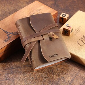 May include: A brown leather journal with a leather strap closure and the name "Martin" embossed on the front. The journal is sitting on a wooden surface with other decorative items.