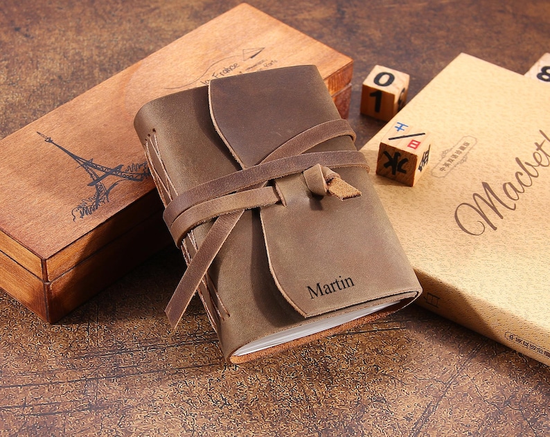 May include: A brown leather journal with a leather strap closure and the name "Martin" embossed on the front. The journal is sitting on a wooden surface with other decorative items.