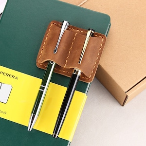 May include: A brown leather pen holder with two pens, one green and one black, attached to a green notebook with the text "PAPERERA squared Notebook".