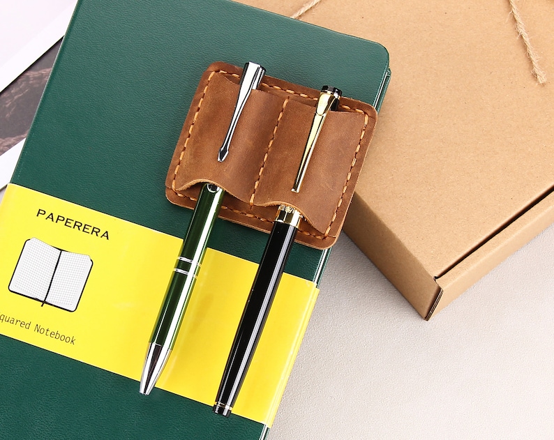 May include: A brown leather pen holder with two pens, one green and one black, attached to a green notebook with the text "PAPERERA squared Notebook".