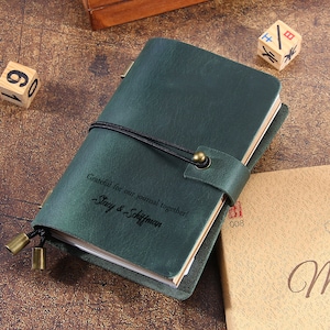 May include: A dark green leather journal with a black elastic closure and a brass clasp. The journal has the inscription "Grateful for our journal together! Stacy & Schiffman." Wooden dice and a tan book are in the background.