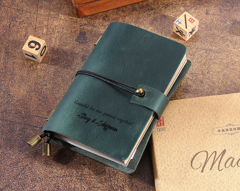 May include: A dark green leather journal with a black elastic closure and a brass clasp. The journal has the inscription "Grateful for our journal together! Stacy & Schiffman." Wooden dice and a tan book are in the background.