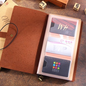 May include: A brown leather journal with a card holder. The journal is open, revealing several cards in the clear plastic sleeves. The cards have various designs and text, including "WH" and "SANFU". A smaller notebook is visible to the left.