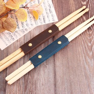 以下が含まれることがあります： 2組の木製ドラムスティック。それぞれ革製のケースに入っています。ケースは茶色と青色。ケースには金色のスナップ留めが付いています。ドラムスティックは楽譜と木製の表面に置かれています。