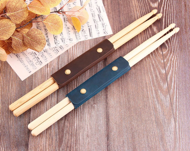 以下が含まれることがあります： 2組の木製ドラムスティック。それぞれ革製のケースに入っています。ケースは茶色と青色。ケースには金色のスナップ留めが付いています。ドラムスティックは楽譜と木製の表面に置かれています。