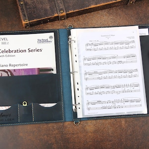 May include: Dark blue leather binder, open to reveal piano sheet music and a music book. The book is part of the "Celebration Series" and is a "Piano Repertoire." A pen lies beside the sheet music. The binder has pockets with the name "Amy's MUSIC LDER".