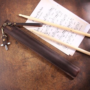Peut inclure: Un &eacute;tui &agrave; baguettes en cuir marron avec un porte-cl&eacute;s en m&eacute;tal. L'&eacute;tui est pos&eacute; sur une surface en bois avec deux baguettes en bois sur une feuille de musique.