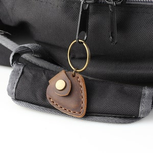 May include: A brown leather key chain with a gold metal ring and a brass snap closure. The key chain is attached to a black backpack.