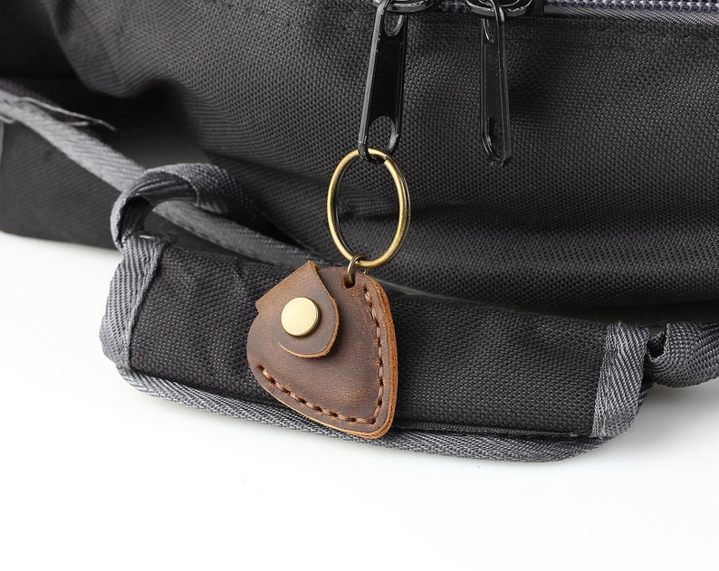 May include: A brown leather key chain with a gold metal ring and a brass snap closure. The key chain is attached to a black backpack.