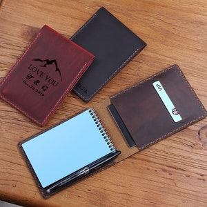 Puede incluir: Tres fundas de cuero para pasaporte en rojo, marrón oscuro y marrón medio. La funda roja presenta un gráfico de montaña con el texto LOVE YOU T & G Dec.25.2019. La funda marrón oscuro tiene las iniciales D.T.K. Una funda está abierta con un cuaderno de espiral azul, un bolígrafo negro y una tarjeta VIP.