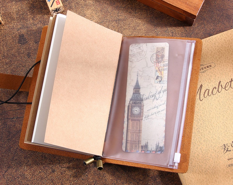 May include: Open brown leather journal with blank pages and a clear pocket holding a bookmark with a Big Ben illustration. The journal is bound with a black cord and has brass-colored metal accents.