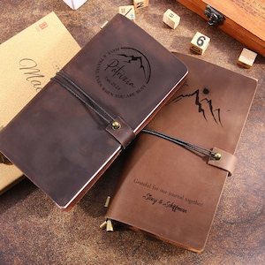 May include: Two brown leather journals with mountain and sun designs. One journal has the name "Patrizia" and the date "Dec. 25.24" engraved. The other journal has the text "Grateful for our journal together! Stacy & Schiffman".
