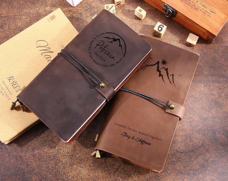 May include: Two brown leather journals with mountain and sun designs. One journal has the name "Patrizia" and the date "Dec. 25.24" engraved. The other journal has the text "Grateful for our journal together! Stacy & Schiffman".