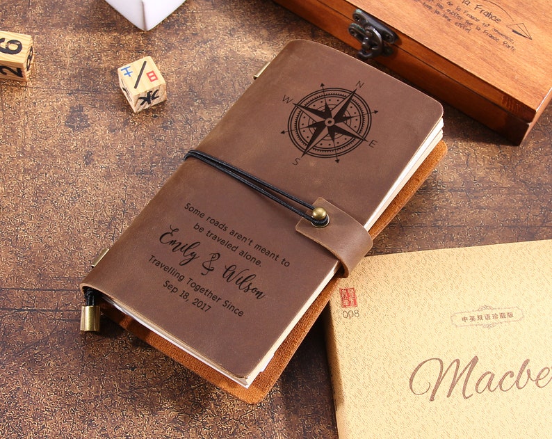 May include: A brown leather journal with a compass rose design and the text "Some roads aren't meant to be traveled alone. Emily & Wilson Travelling Together Since Sep 18, 2017." The journal is secured with a black elastic band.