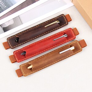 May include: Three leather pen holders with elastic bands. The holders are brown, red, and tan. Each holder has a pen inside.