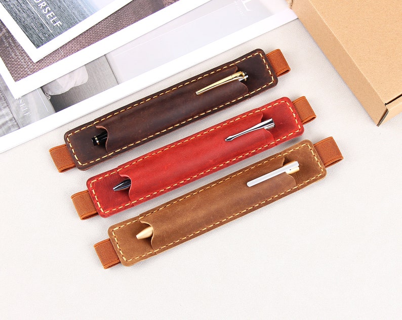 May include: Three leather pen holders with elastic bands. The holders are brown, red, and tan. Each holder has a pen inside.