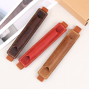 May include: Three leather pen holders in brown, red, and tan with elastic bands. The holders are stitched with a contrasting thread.