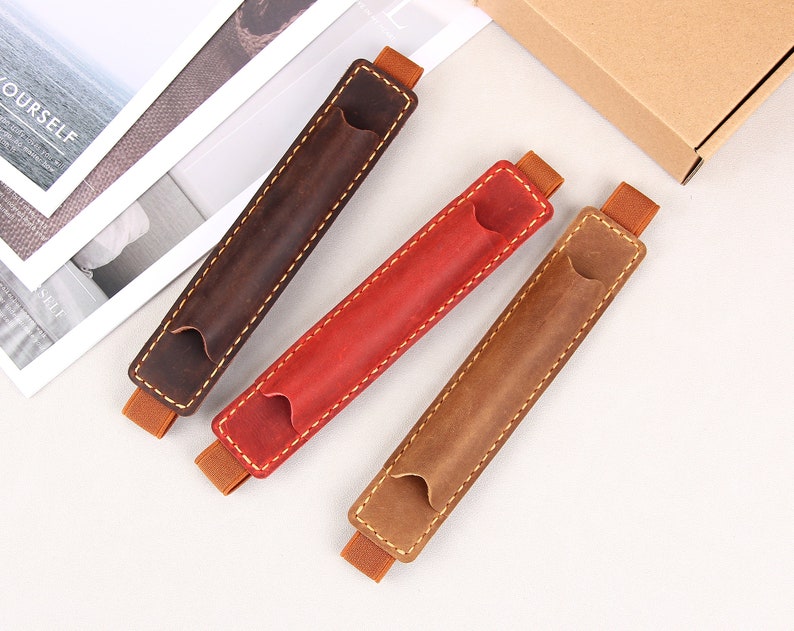 May include: Three leather pen holders in brown, red, and tan with elastic bands. The holders are stitched with a contrasting thread.