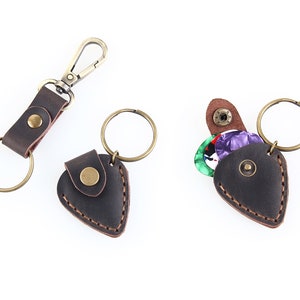 May include: A brown leather guitar pick holder with a key ring and a snap closure. The holder is shaped like a guitar pick and has a small pocket for storing picks. The holder is shown with a few colorful guitar picks inside.
