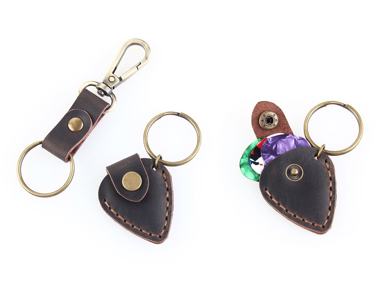 May include: A brown leather guitar pick holder with a key ring and a snap closure. The holder is shaped like a guitar pick and has a small pocket for storing picks. The holder is shown with a few colorful guitar picks inside.
