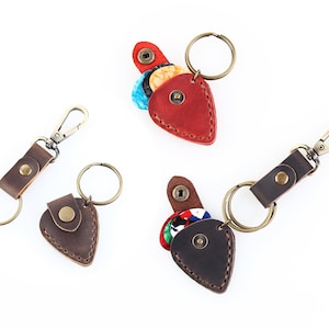 May include: Three leather guitar pick holders with keychains. The holders are shaped like guitar picks and have a flap that opens to reveal a space for picks. One holder is red, one is brown, and one is dark brown.