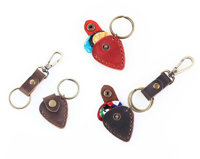May include: Three leather guitar pick holders with keychains. The holders are shaped like guitar picks and have a flap that opens to reveal a space for picks. One holder is red, one is brown, and one is dark brown.
