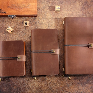 May include: Three brown leather journals of varying sizes, secured with black elastic and leather straps. The journals are arranged on a wooden surface, with a wooden box and dice in the background. The journals have a classic, vintage aesthetic.