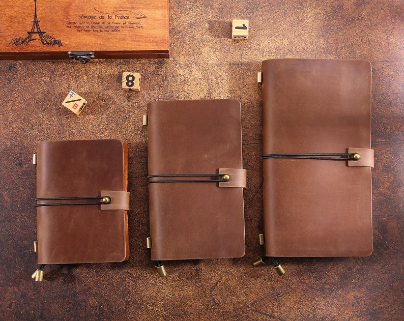 May include: Three brown leather journals of varying sizes, secured with black elastic and leather straps. The journals are arranged on a wooden surface, with a wooden box and dice in the background. The journals have a classic, vintage aesthetic.