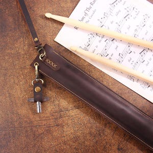 Peut inclure: Une sangle en cuir marron avec un porte-cl&eacute;s et une petite &eacute;tiquette m&eacute;tallique portant le mot "XXXX". La sangle est pos&eacute;e sur une surface en bois avec une feuille de musique en dessous.