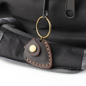 May include: A brown leather guitar pick holder with a brass ring attached to a key chain. The holder is attached to a black canvas bag.