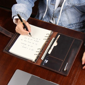 May include: A brown leather bound journal with a pen and a card holder. The journal has a ring binder and is open to a blank page with handwritten notes.