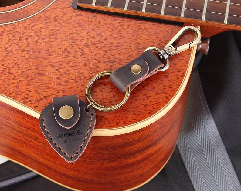 May include: A brown leather guitar pick keychain with a metal ring and a silver-toned clasp. The pick is shaped like a heart and has the text "James S. 10.11.2024" engraved on it.