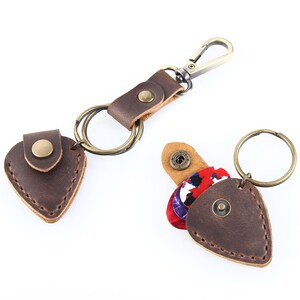 May include: Two brown leather guitar pick holders with key rings. One holder has a single pick, the other has multiple picks. Both holders have a brown leather strap with a metal clasp.