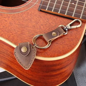 May include: A brown leather guitar pick keychain with a metal ring and a silver clasp. The keychain is attached to an acoustic guitar.