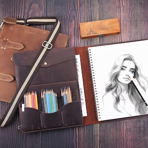 May include: A brown leather art portfolio with a shoulder strap, open to reveal a sketchbook with a pencil drawing of a woman. A pencil case and a set of colored pencils are also visible. The portfolio is on a wooden surface.