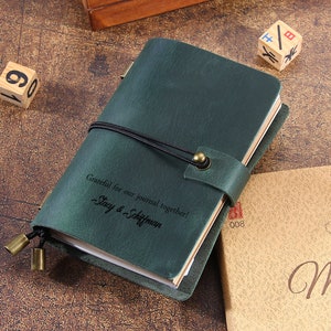 May include: A green leather journal with a black elastic closure and a brass clasp. The journal has the text "Grateful for our journal together! Stacy & Schiffman" engraved on the front cover.