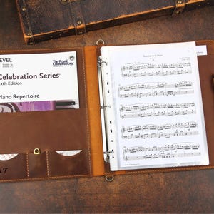 Peut inclure: Un classeur en cuir marron avec un protège-feuilles en plastique transparent contenant de la musique. Le classeur a une poche sur le devant avec le texte "The Royal Conservatory Celebration Series Sixth Edition Piano Repertoire" et les initiales "B&T" en bas à gauche.