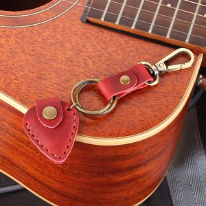 May include: A red leather guitar pick holder attached to a silver key chain with a gold ring. The keychain is hanging on a brown acoustic guitar.