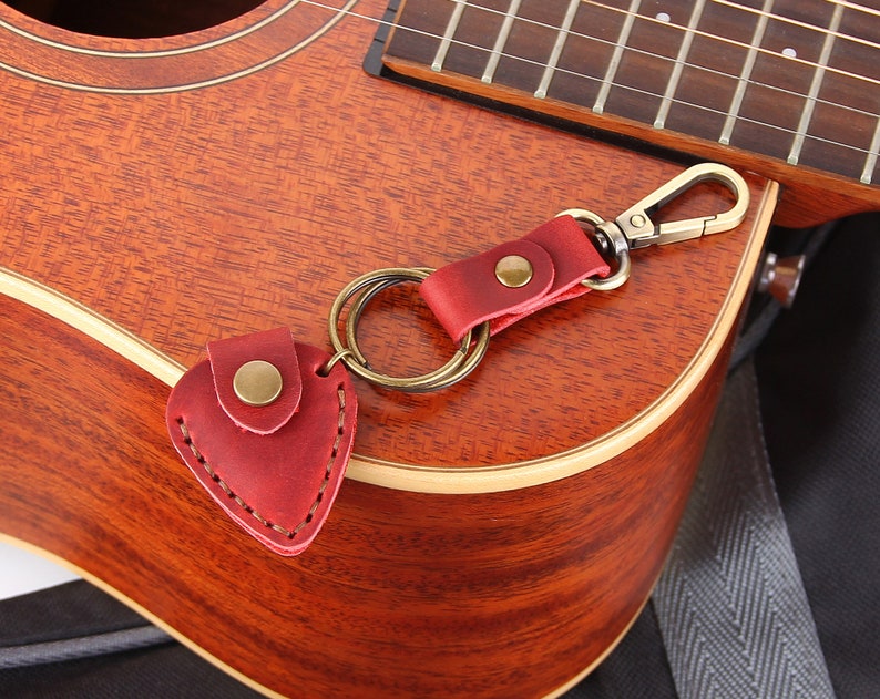 May include: A red leather guitar pick holder attached to a silver key chain with a gold ring. The keychain is hanging on a brown acoustic guitar.