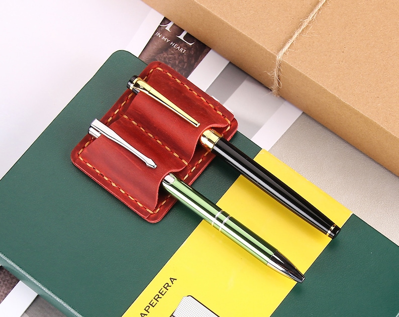 May include: A red leather pen holder with two slots for pens. The holder has a stitched edge and is sitting on a green and yellow notebook.