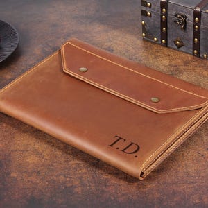 May include: A brown leather document folder featuring a flap closure secured with two brass-colored metal snaps. The front of the folder is embossed with the initials T.D. in a dark, serif font, and the edges are finished with visible stitching.