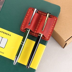 May include: A red leather pen holder with two slots for pens. The holder is attached to a green and yellow notebook with the text "PAPERERA Squared Notebook".