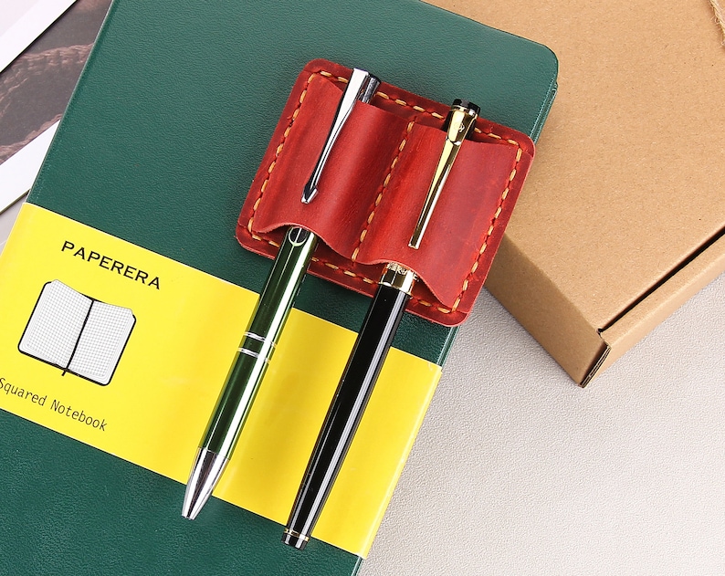 May include: A red leather pen holder with two slots for pens. The holder is attached to a green and yellow notebook with the text "PAPERERA Squared Notebook".