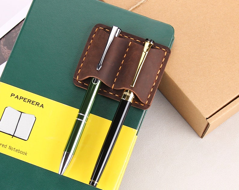 May include: A brown leather pen holder with two slots for pens. The holder is attached to a green notebook with the word "PAPERERA" printed on the front. The notebook has a yellow stripe with the words "red Notebook" printed on it.