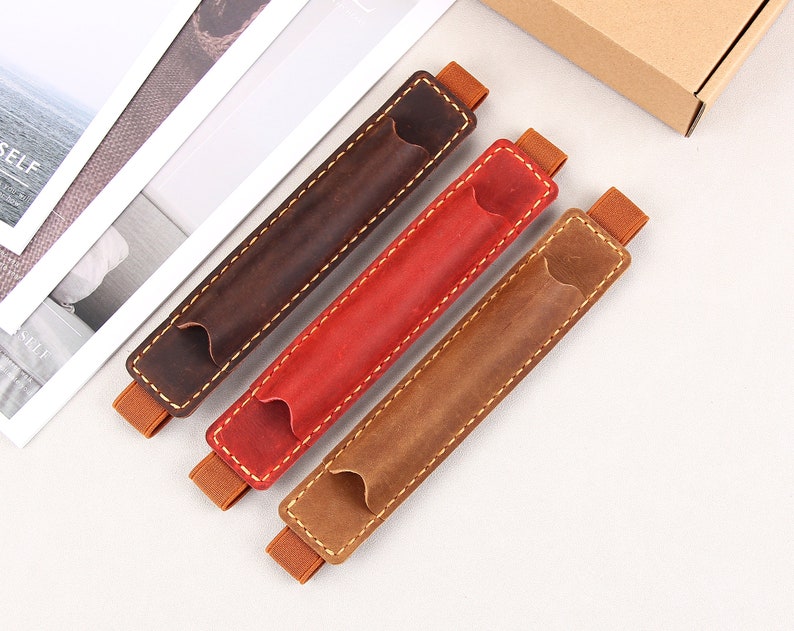 May include: Three leather pen holders in brown, red, and tan. Each holder has a stitched seam and an elastic band at each end.