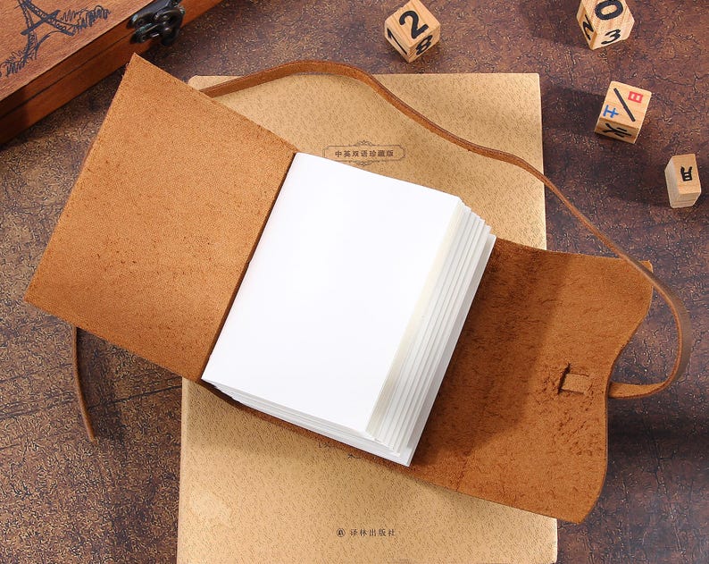 May include: A brown leather bound journal with blank pages. The journal is open and lying on a brown surface. The journal has a strap closure and a decorative metal clasp.