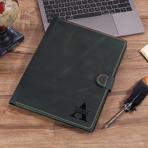 May include: A dark green leather notebook cover featuring a snap closure and yellow contrast stitching. The front is personalized with a large black letter A and the name Andrew printed across the center of the letter in a horizontal line.