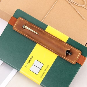 May include: A green leather-bound notebook with a yellow label that says "PAPERERA Squared Notebook". A brown leather pen holder with a gold pen is attached to the notebook.