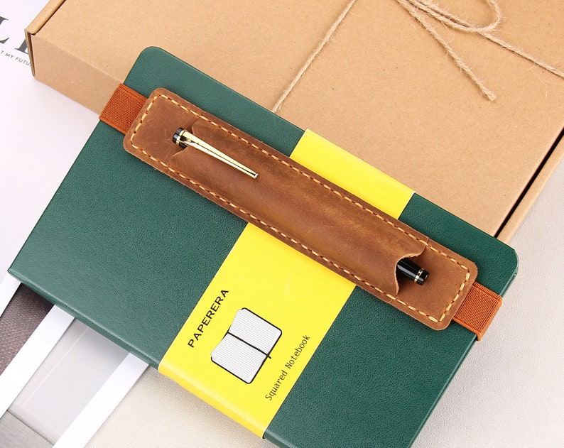 May include: A green leather-bound notebook with a yellow label that says "PAPERERA Squared Notebook". A brown leather pen holder with a gold pen is attached to the notebook.